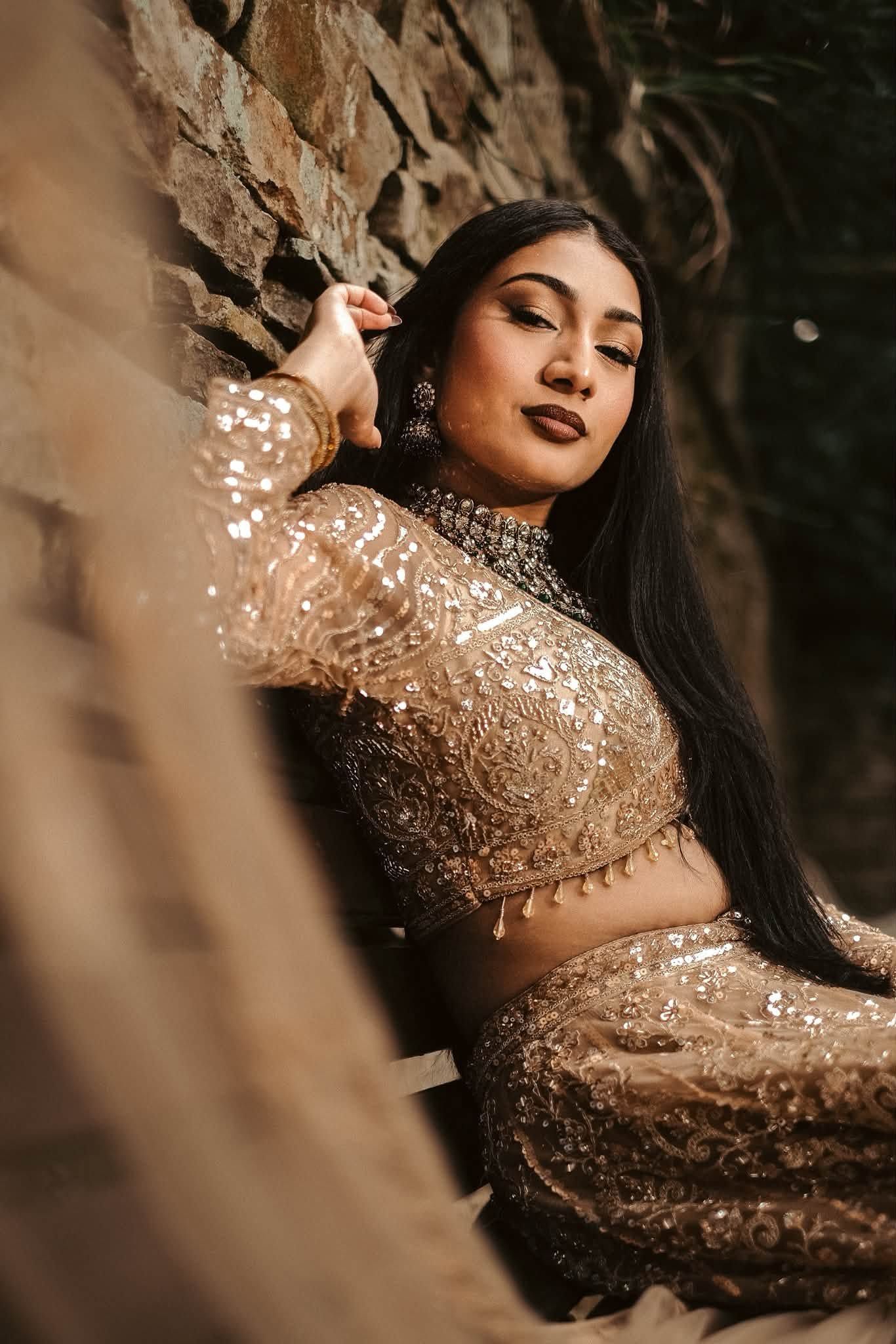 A woman in a gold embroidered lehenga poses gracefully against a rustic stone wall.