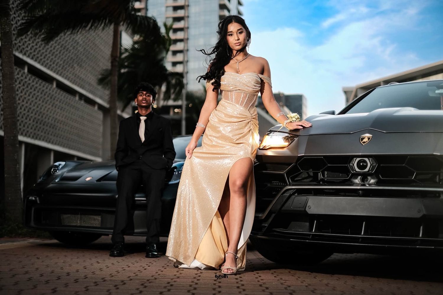 Young woman in gold slit gown leaning on Lamborghini with suited date behind her.