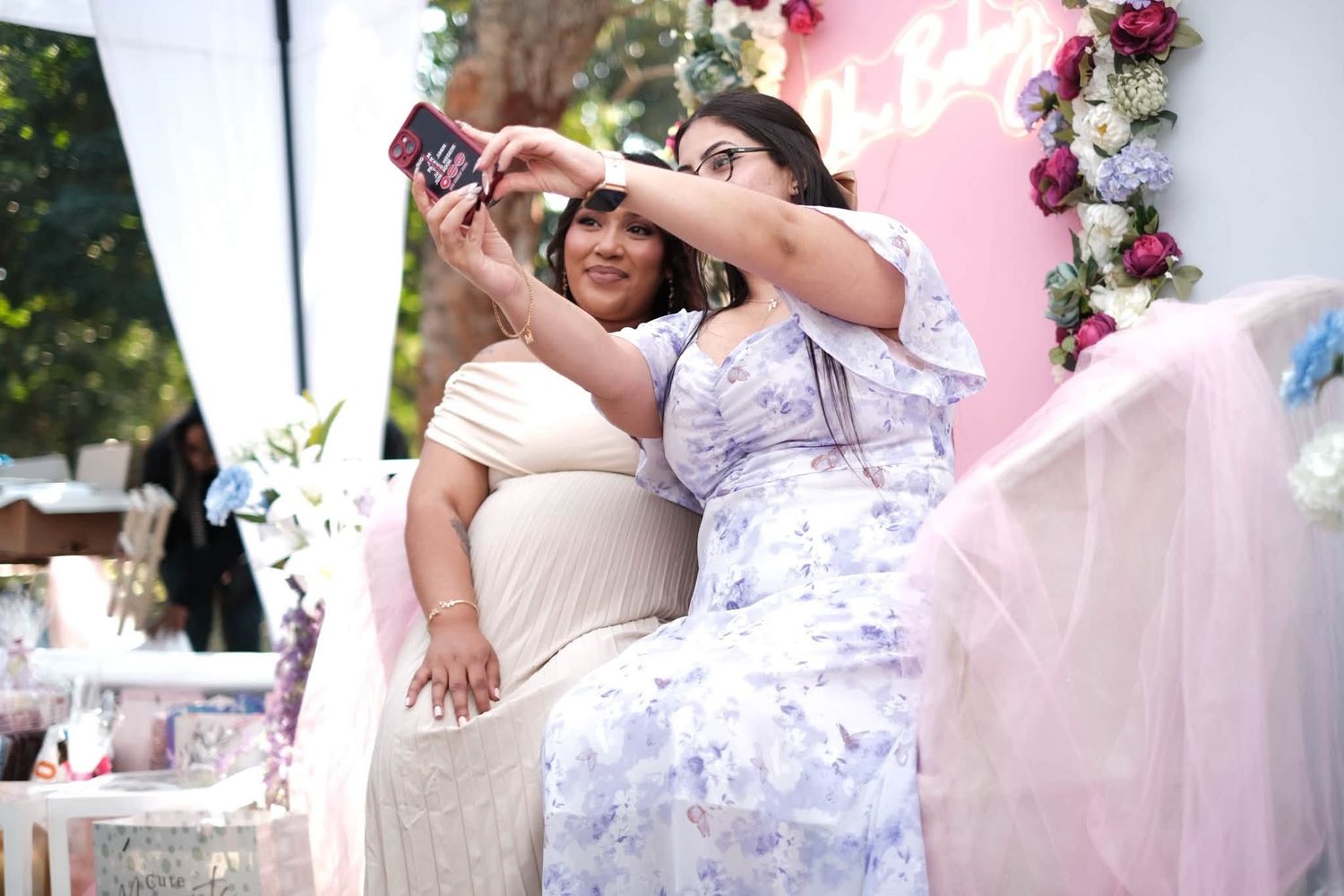 Two women in floral dresses take a selfie at an outdoor baby shower with pink floral decorations.