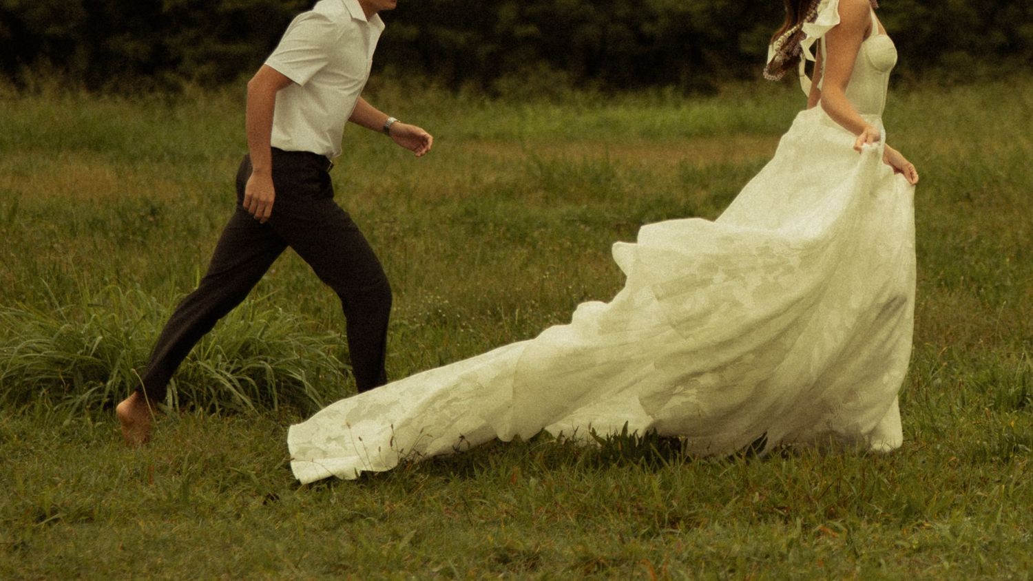 Bride in flowing white gown running through green meadow with groom following behind.