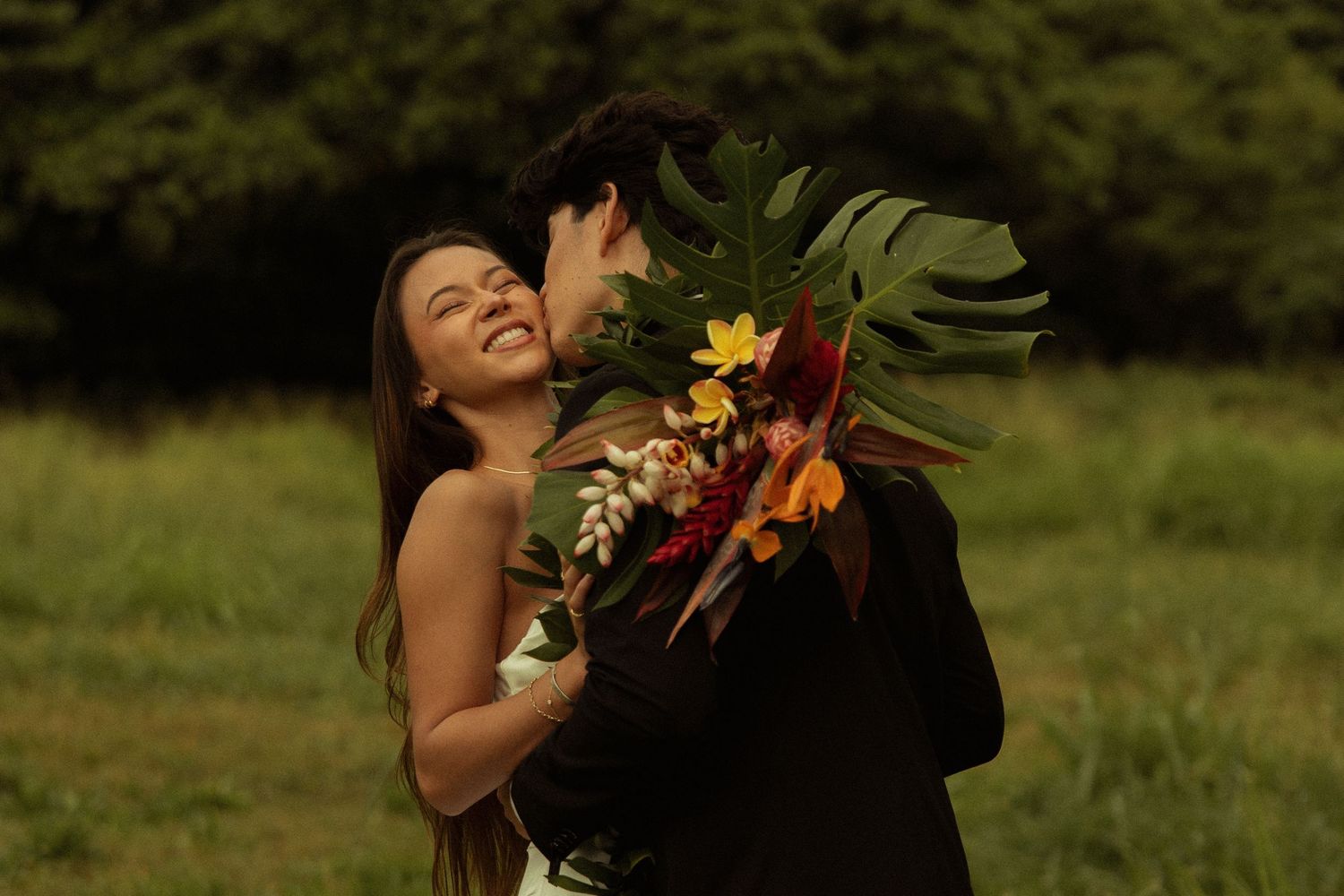 Smiling bride hugging groom holding a tropical wedding bouquet in a green field.