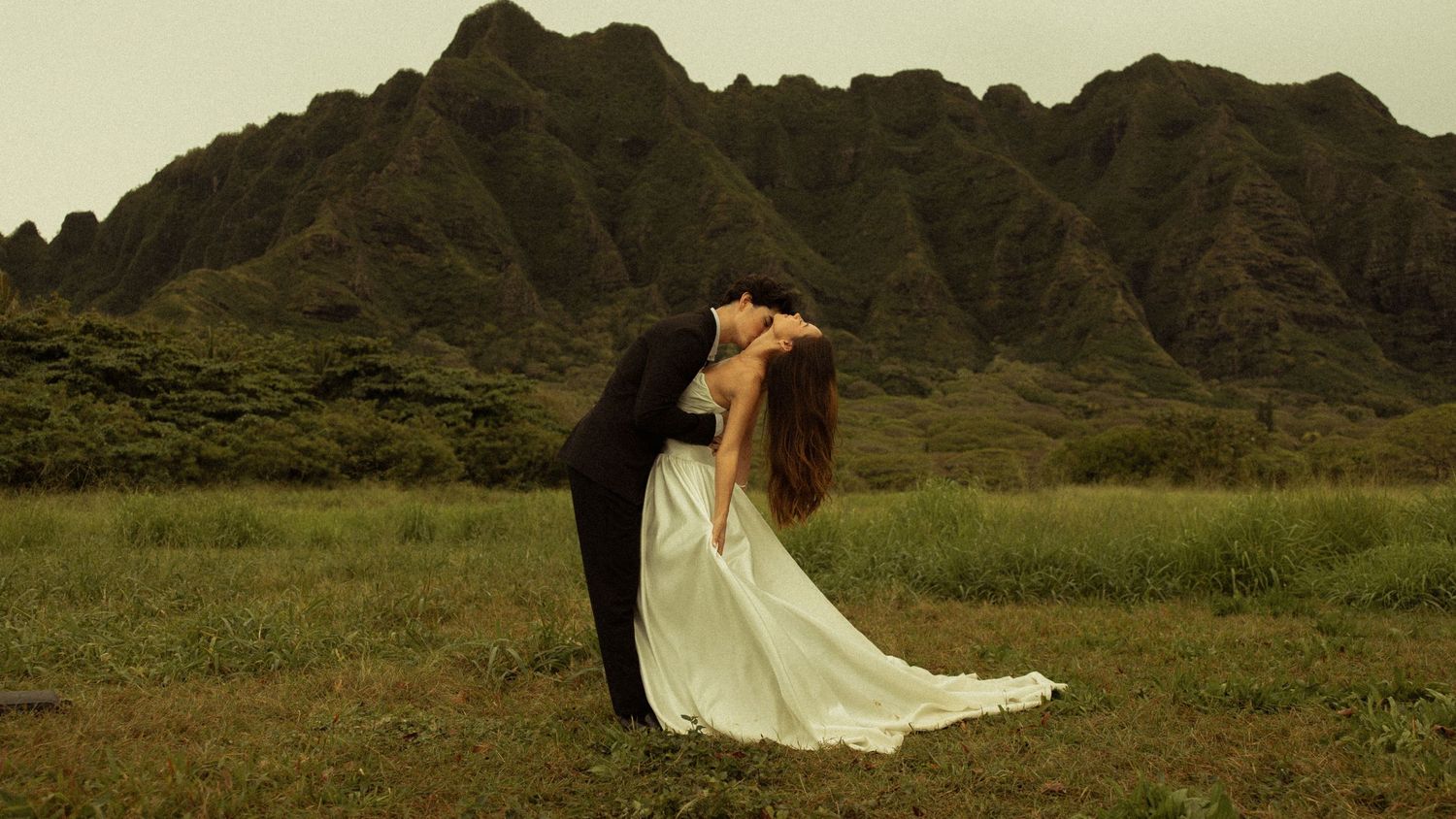 Groom dips bride in white dress for a romantic kiss in front of lush mountain backdrop.