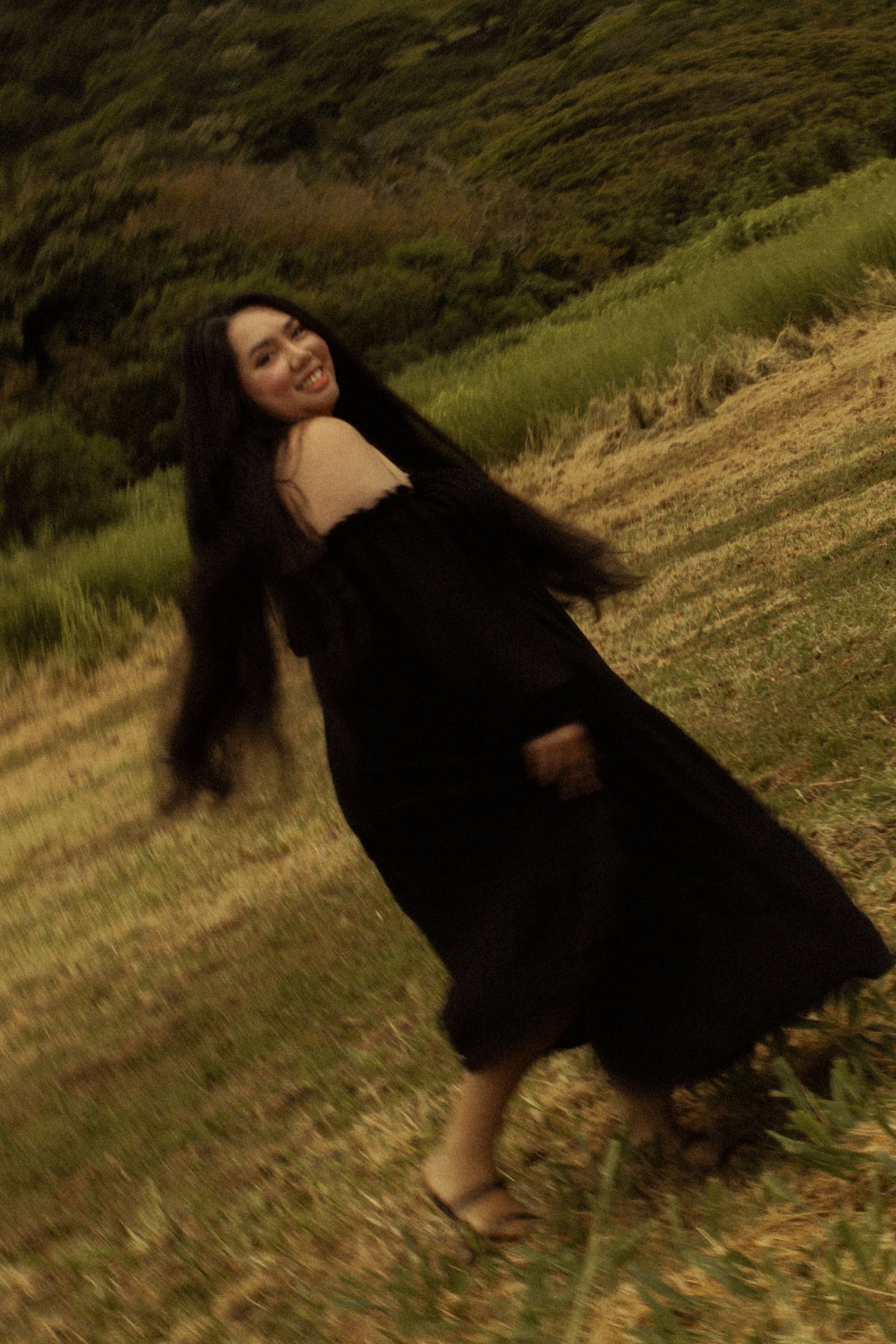 Smiling woman in black dress spins on a hillside with wind blowing her long dark hair.