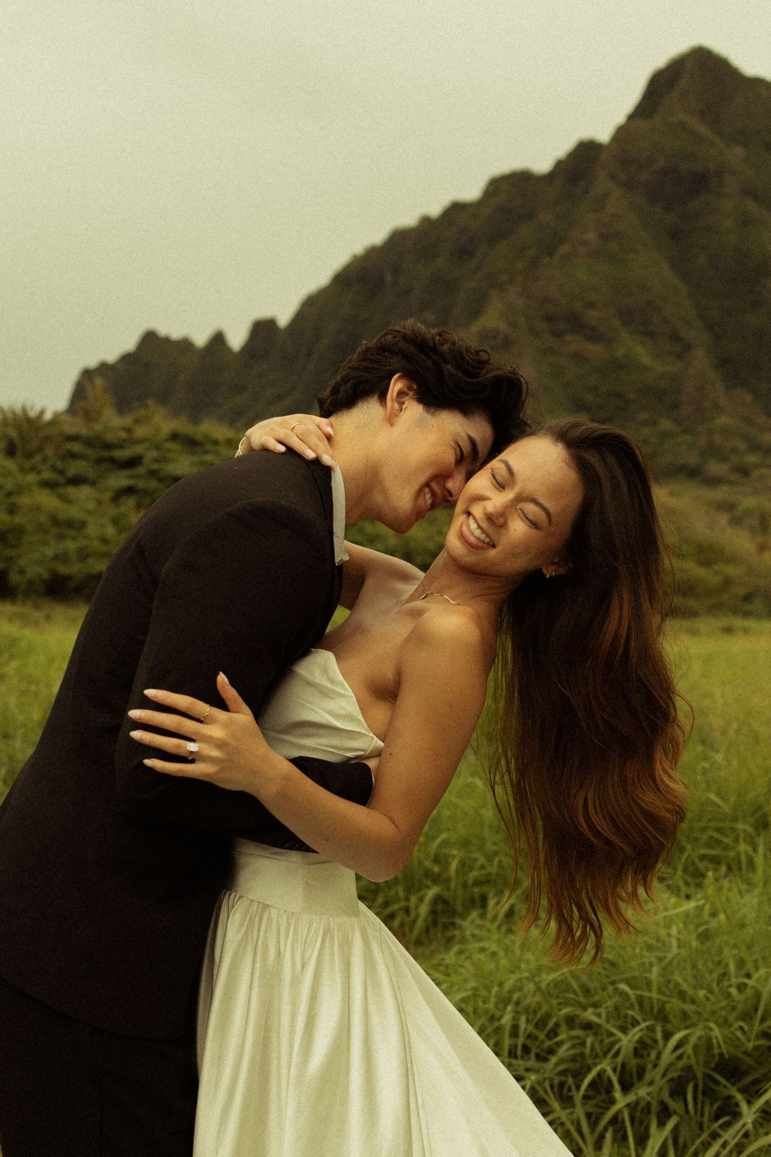 Laughing bride in white dress embraces groom in black suit with green mountains behind them.