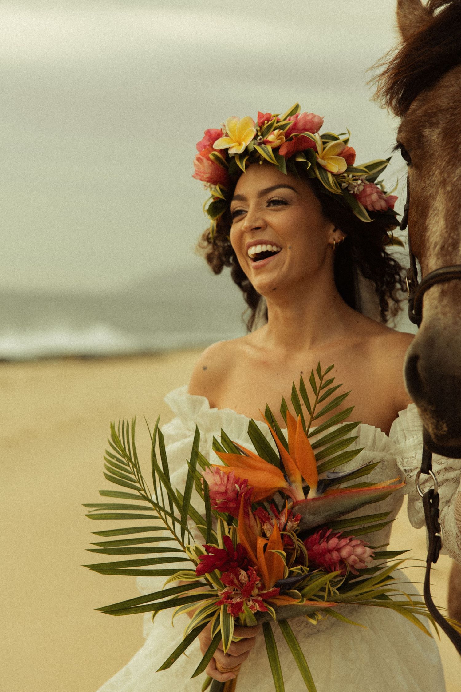 Joyful bride in floral crown laughs while holding tropical bouquet beside horse on beach.