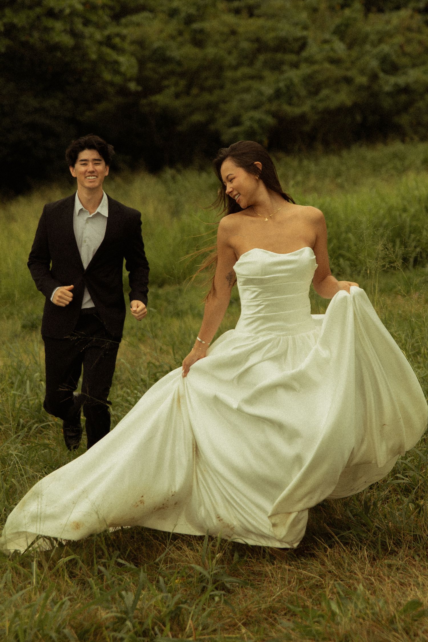 Bride in strapless white ballgown running through green field with smiling groom following.