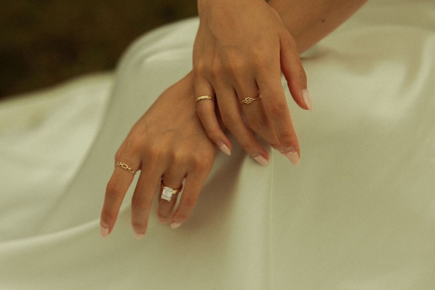 Close-up of bride's hands adorned with gold rings resting on white wedding dress fabric.