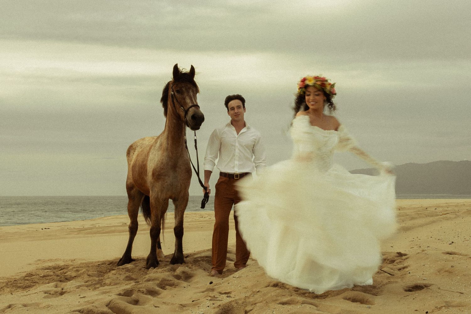 Bride and groom posing with brown horse on sandy beach under cloudy sky.