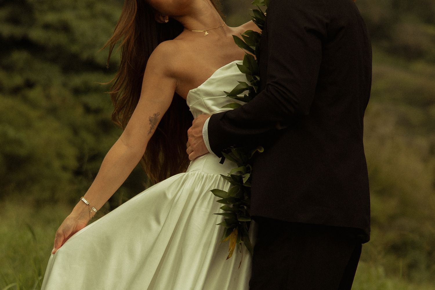 Groom in black suit embracing bride in strapless white gown with tropical greenery.