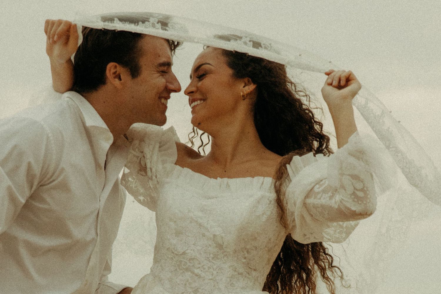 Happy couple in white wedding attire laughing under a flowing veil on a beach.