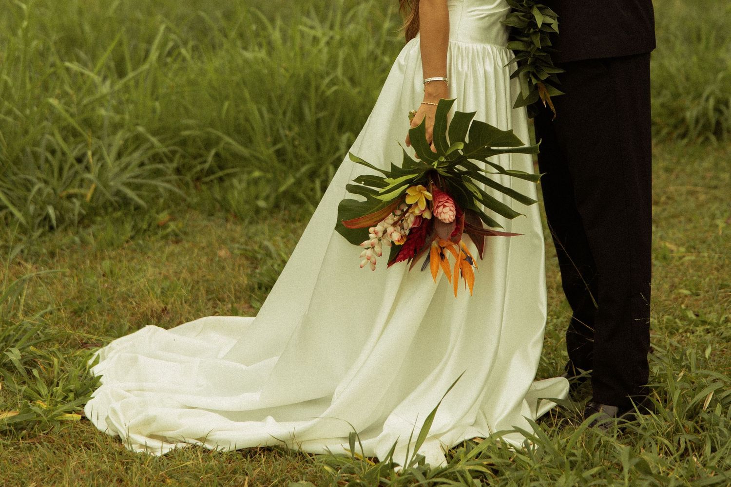 Close-up of bride in flowing white gown holding a tropical bouquet with monstera leaves.