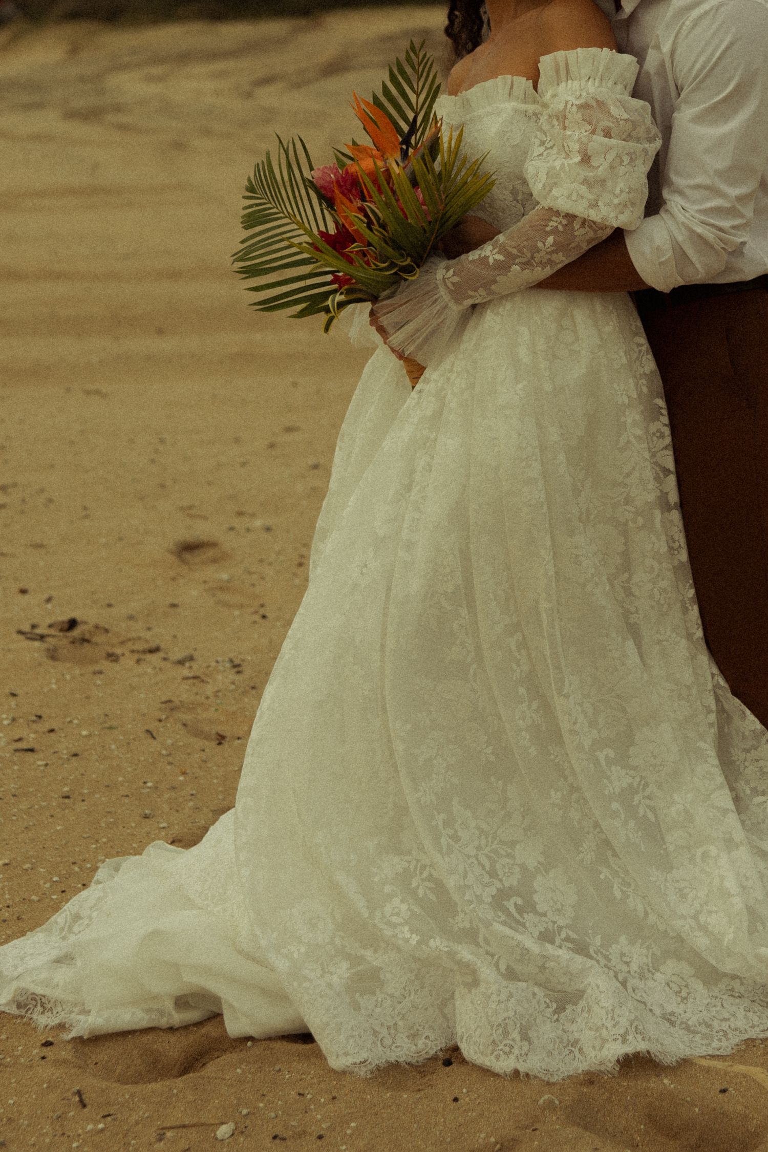 Bride in white lace strapless gown holding colorful tropical bouquet on sandy beach.