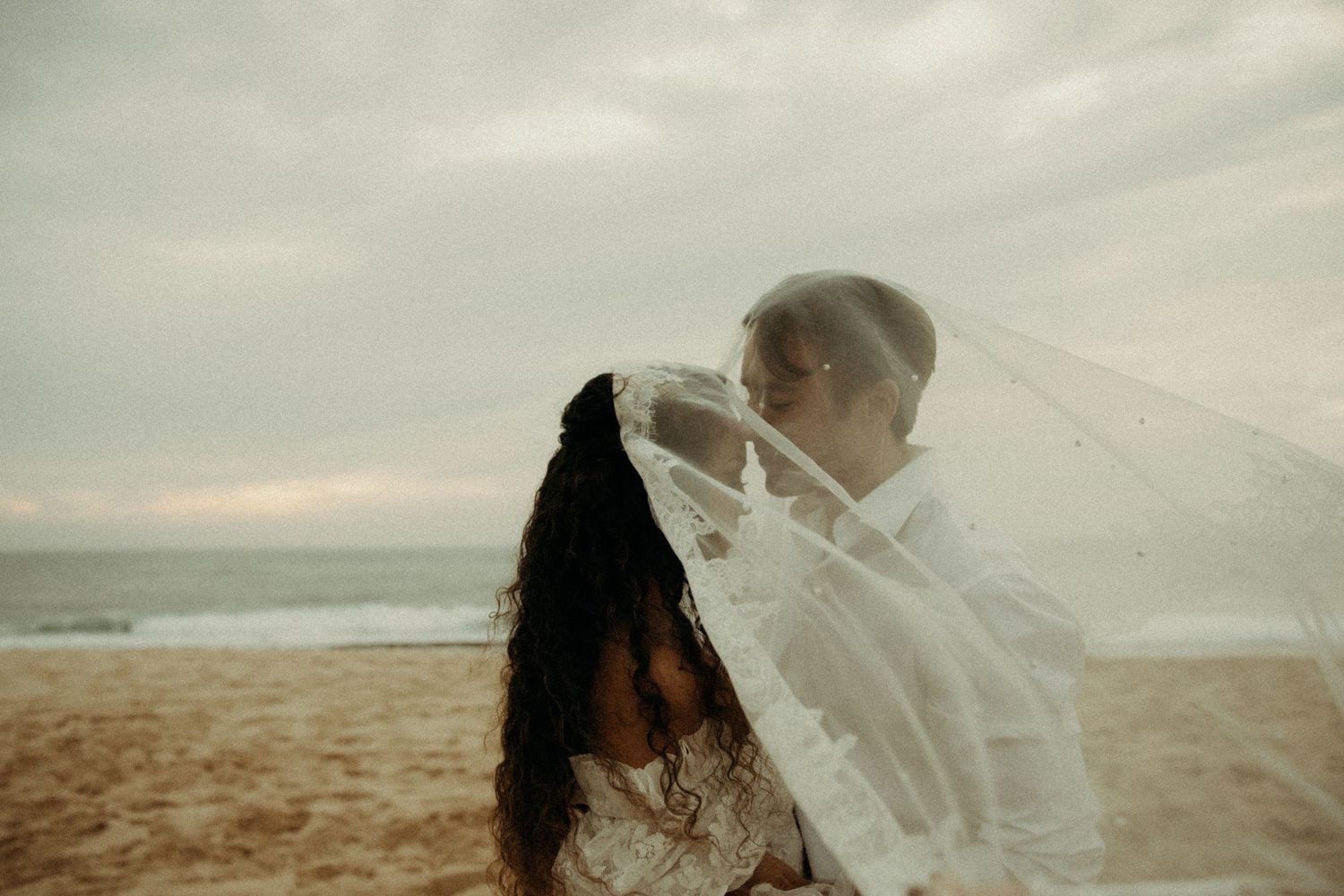 Couple sharing a tender kiss on the beach, bride's veil billowing softly in the ocean breeze.