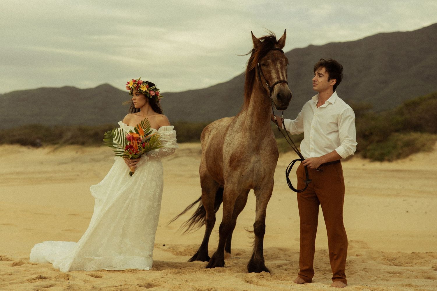 Bride with tropical floral crown and bouquet stands beside brown horse with groom on desert beach.