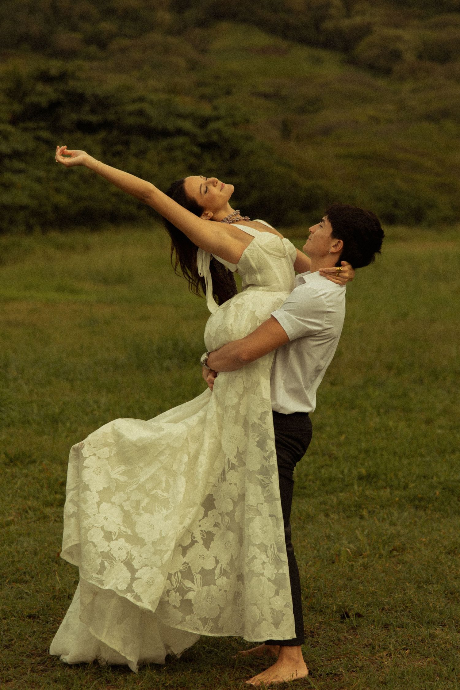 Couple dancing outdoors, woman in white lace dress stretching arms back while man embraces her.
