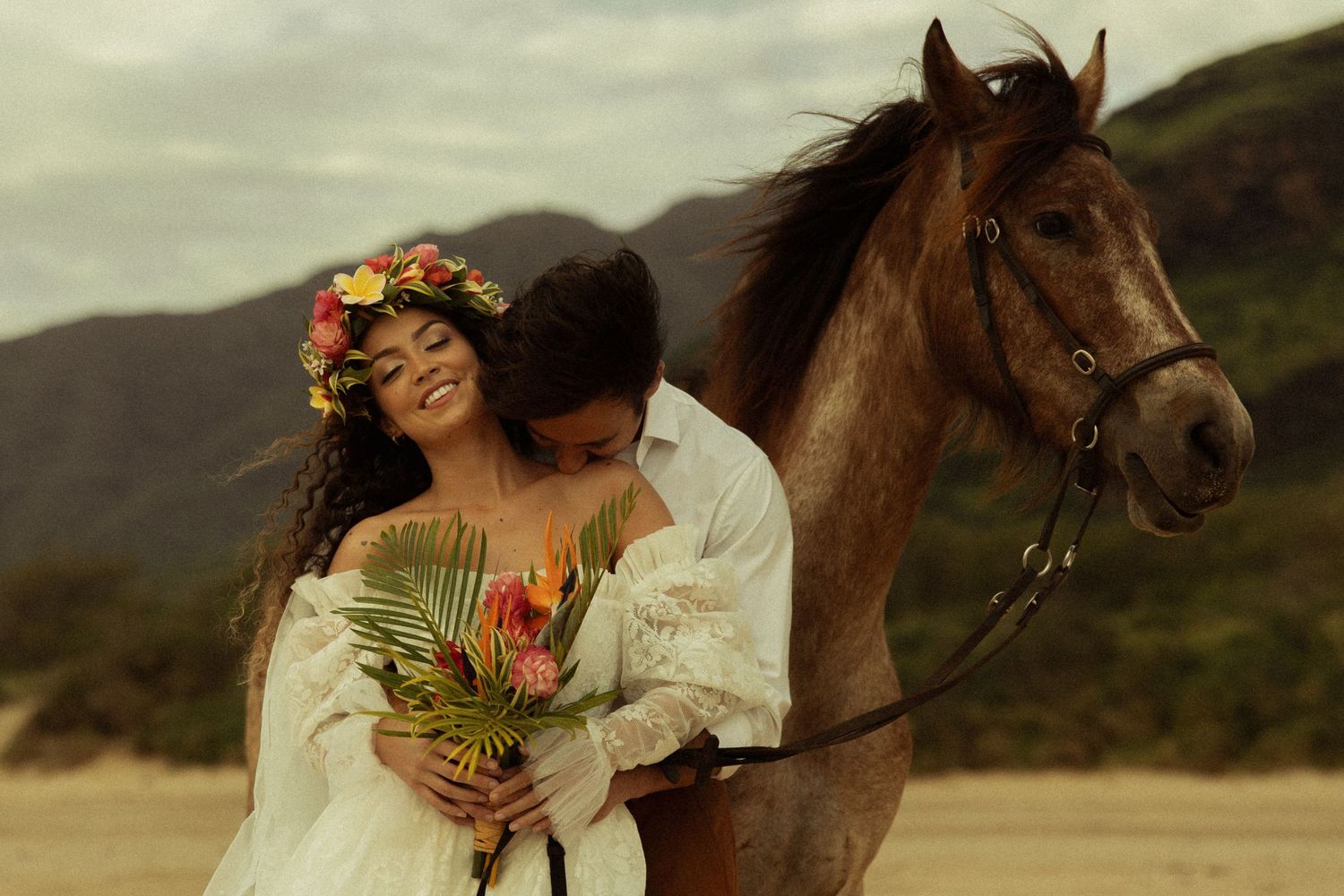 Groom embraces smiling bride holding tropical bouquet beside brown horse on sandy beach.