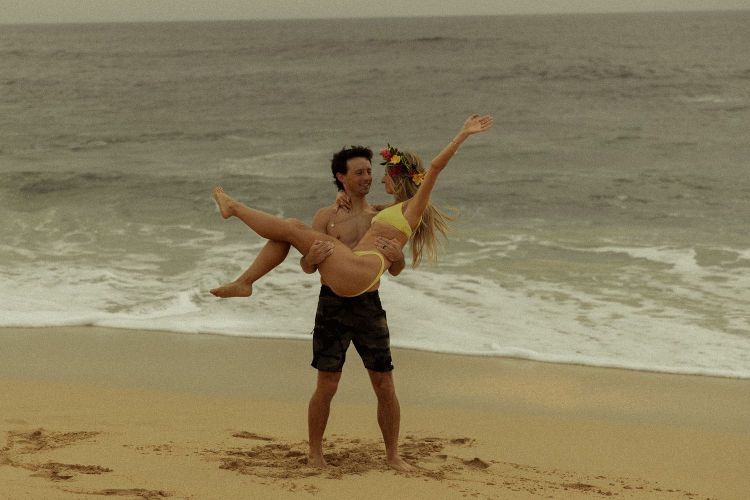 Groom lifting smiling bride on a sandy beach with ocean waves behind them.