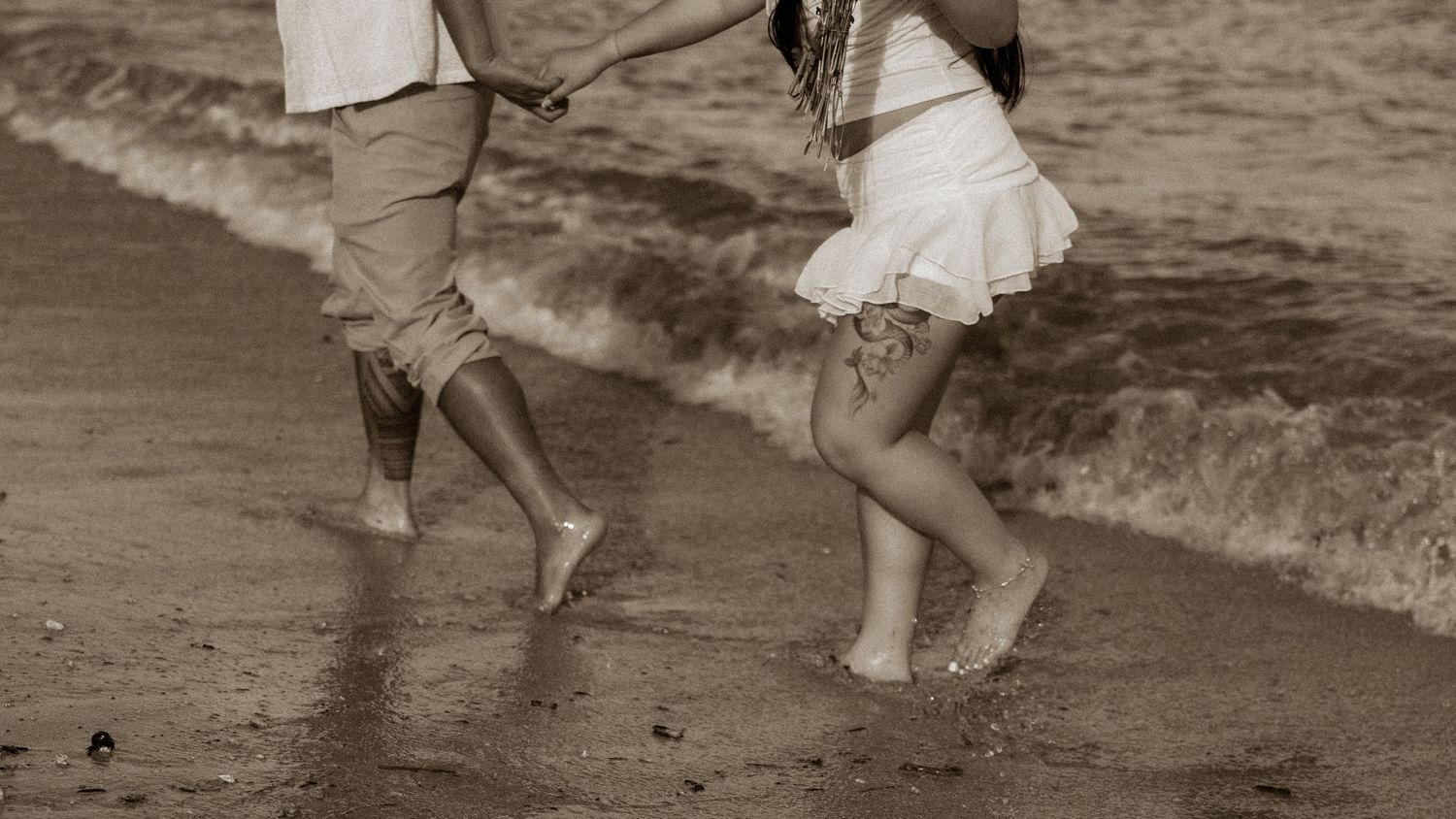 Couple holding hands and running barefoot along a sandy beach shore in sepia tone.