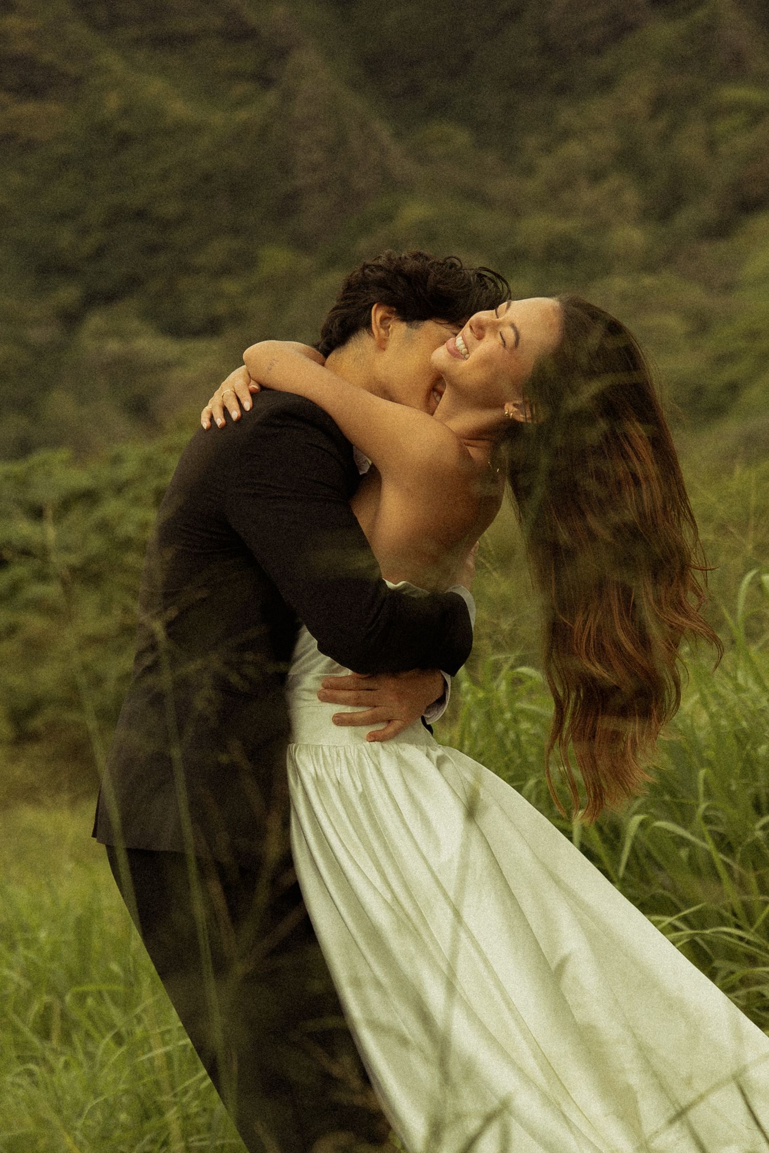 Groom lifting bride in white dress as she laughs joyfully against lush green mountains.