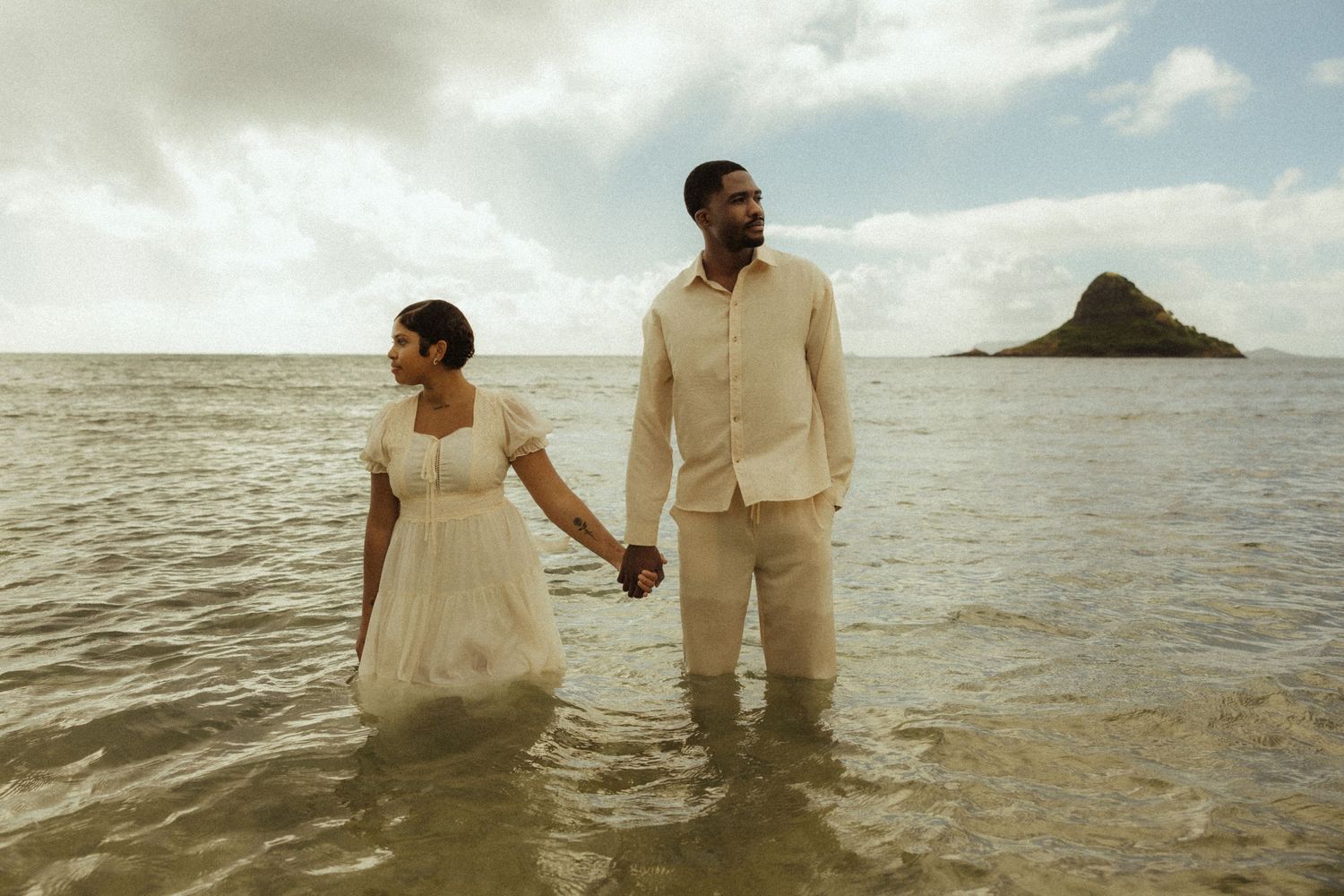 Newlyweds in white wedding clothes holding hands standing in calm ocean water near tropical island.