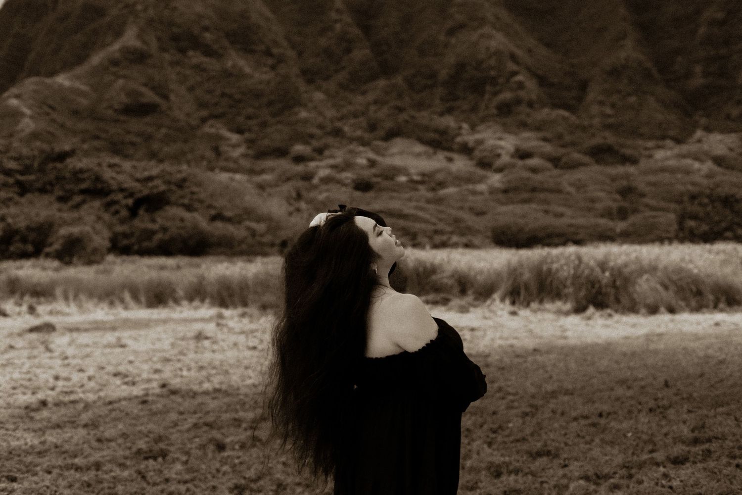 Woman with long dark hair poses dramatically in a field with lush green mountains in the background.