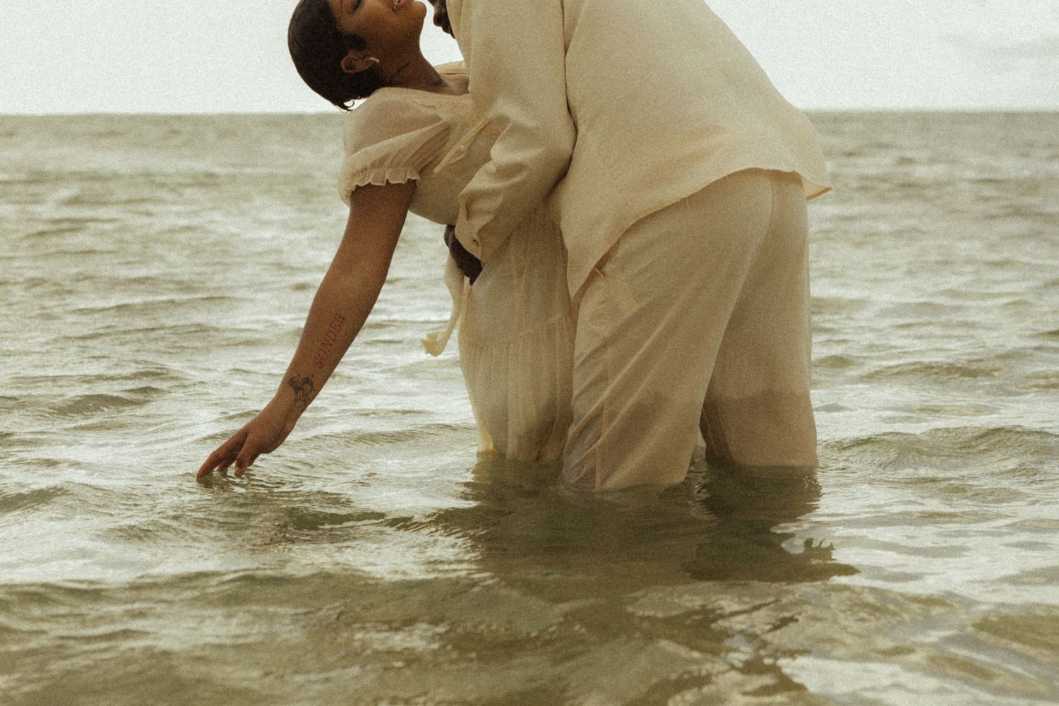Couple standing in shallow ocean water in white outfits, reaching down to touch the sea surface.