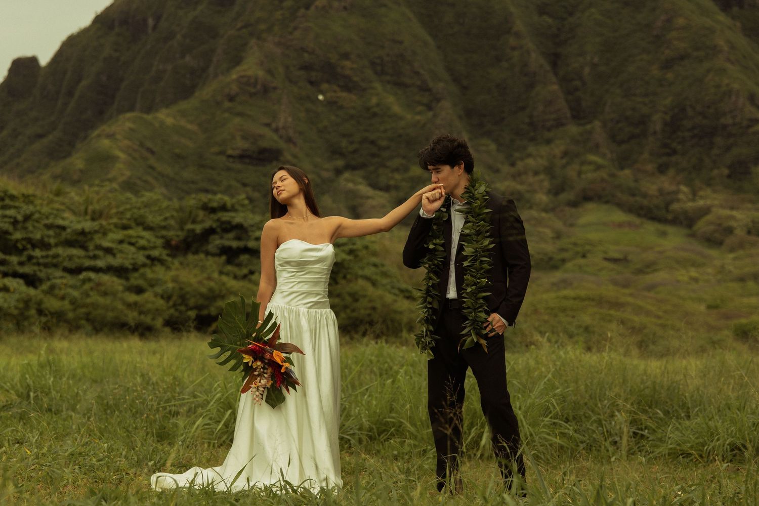 Bride in white gown and groom in black suit posing in a lush Hawaiian valley landscape.