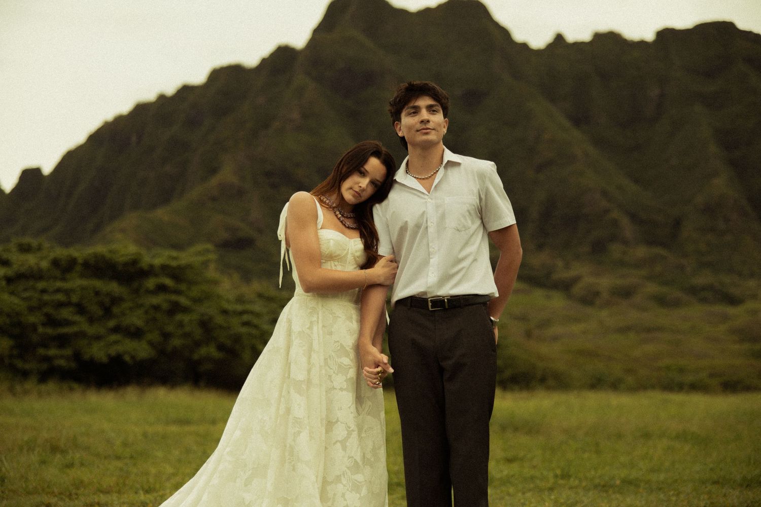 Couple in wedding attire holding hands in a lush green field with dramatic mountains in the background.