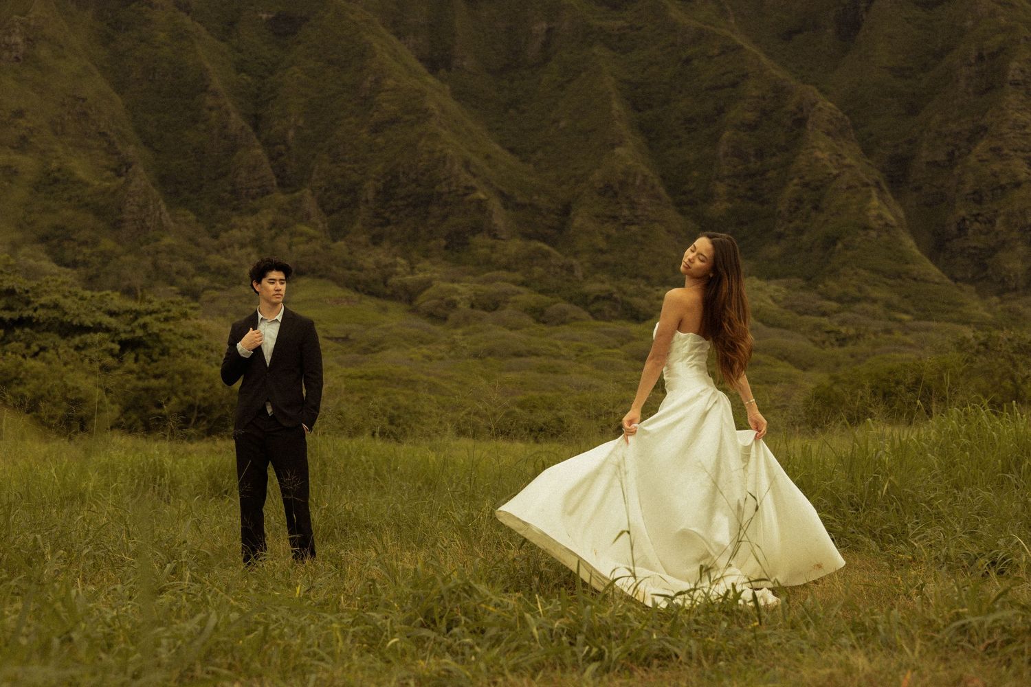 Wedding couple in formal attire pose in a lush green valley with mountain backdrop.