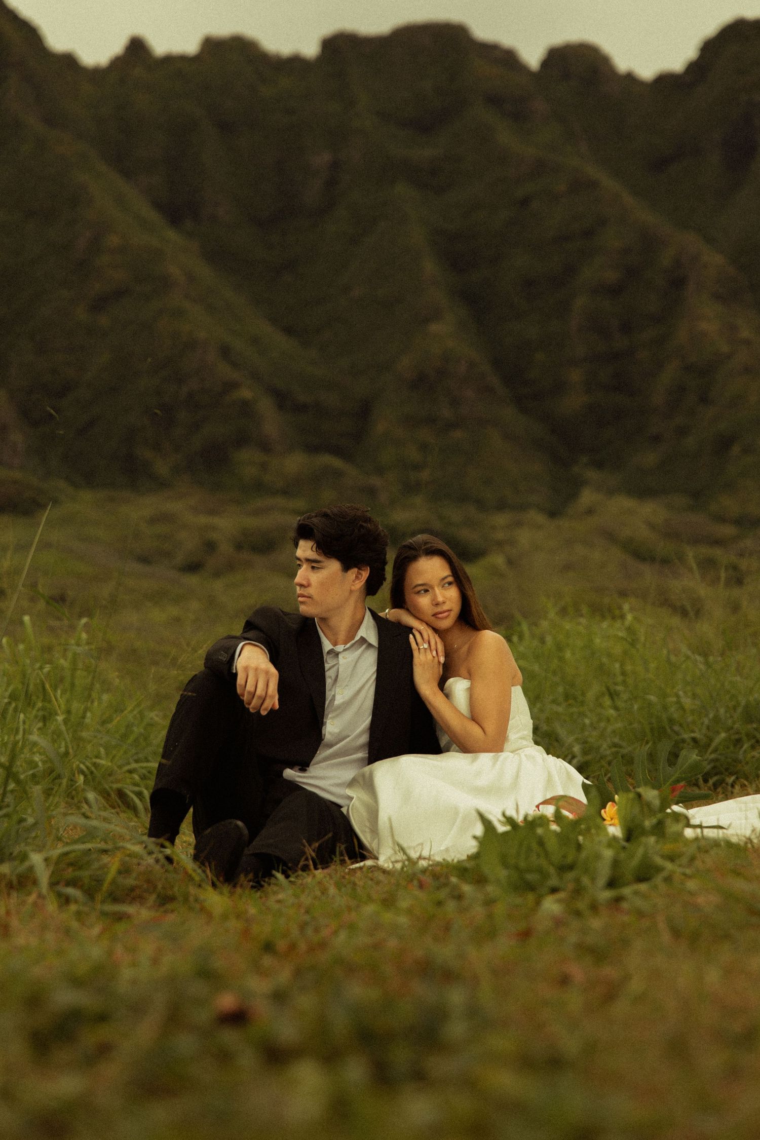 Couple sits together in grassy field gazing into distance with dramatic mountain range backdrop.