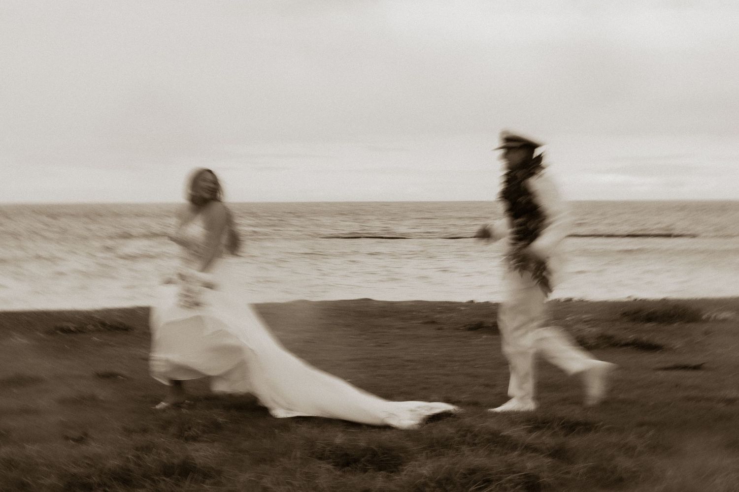 Bride and groom running along a coastal shoreline in a dreamy sepia-toned motion-blurred photograph.