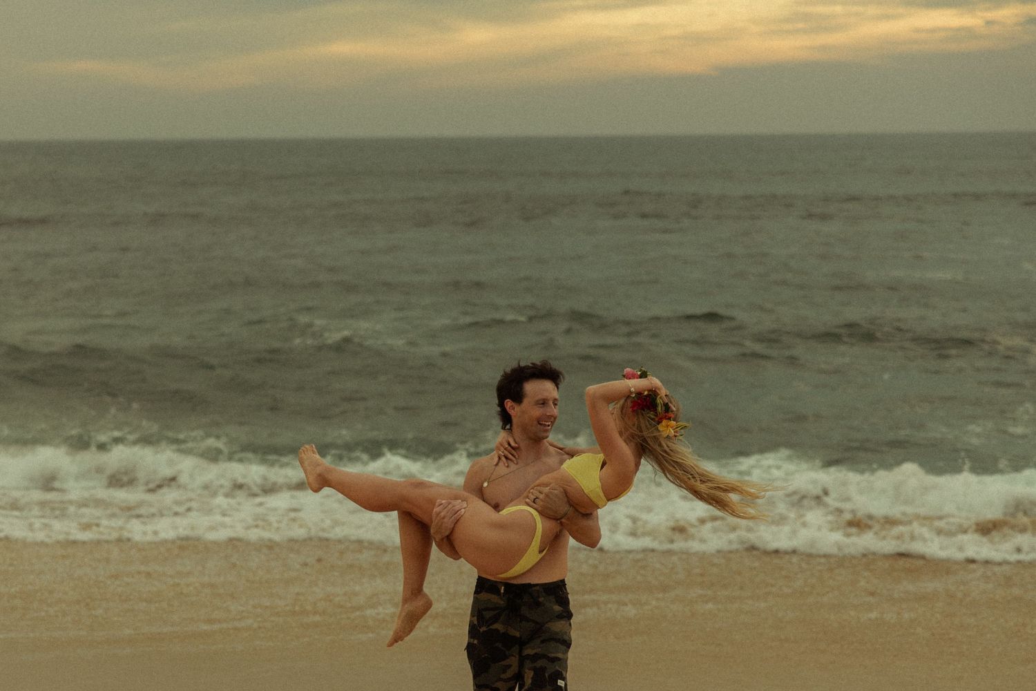 Man carrying woman in yellow bikini on a beach at sunset with ocean waves behind them.