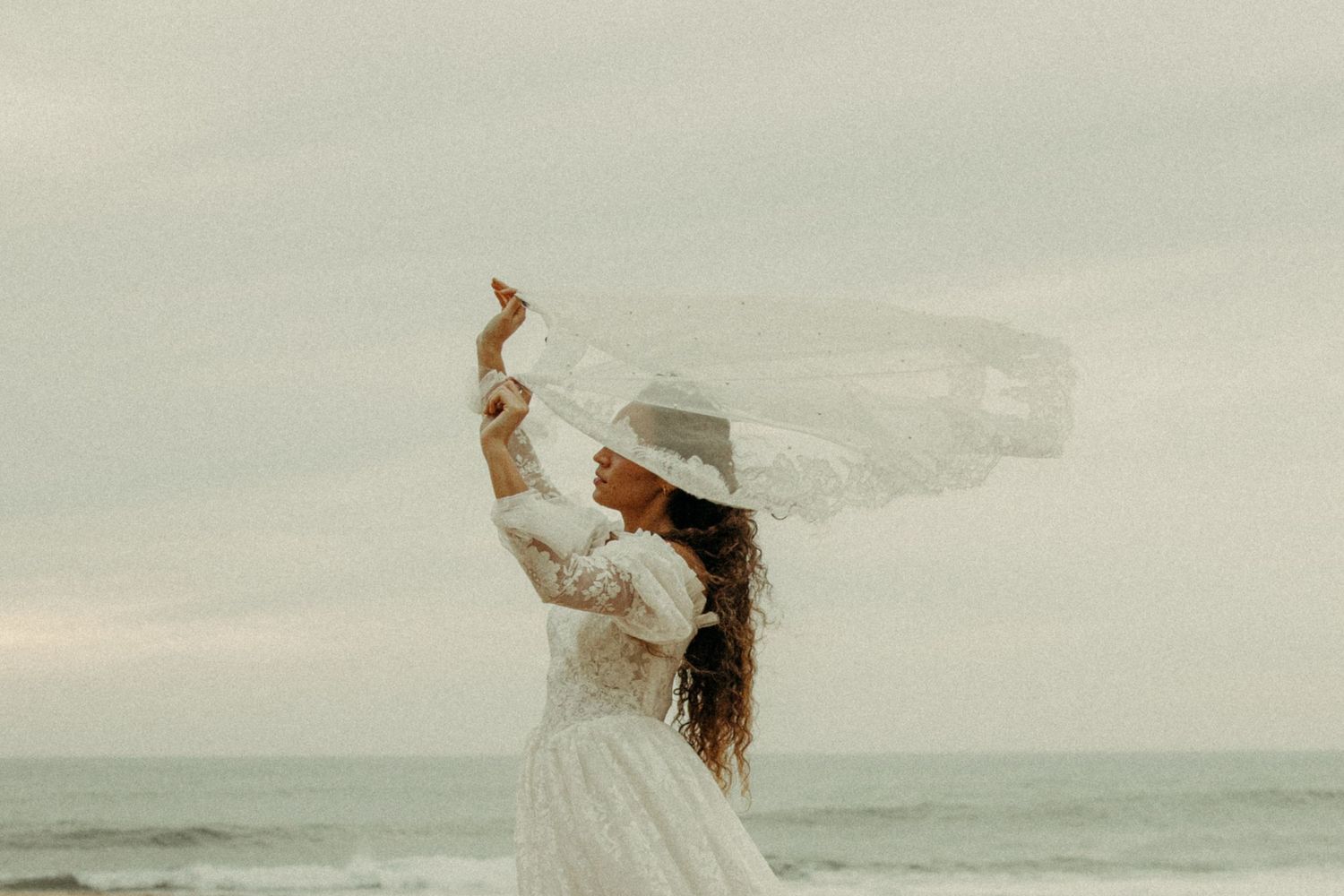 Bride in white lace gown and hat poses dramatically as veil blows in ocean breeze.