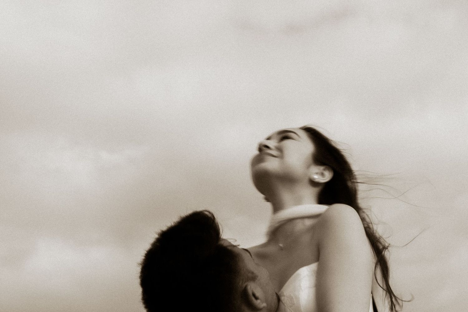 Sepia toned romantic couple photo with woman tilting head back smiling under cloudy sky.
