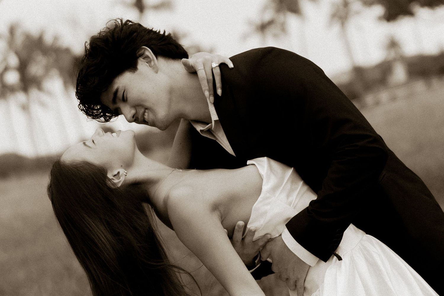 Sepia-toned romantic photo of bride and groom almost kissing under palm trees.
