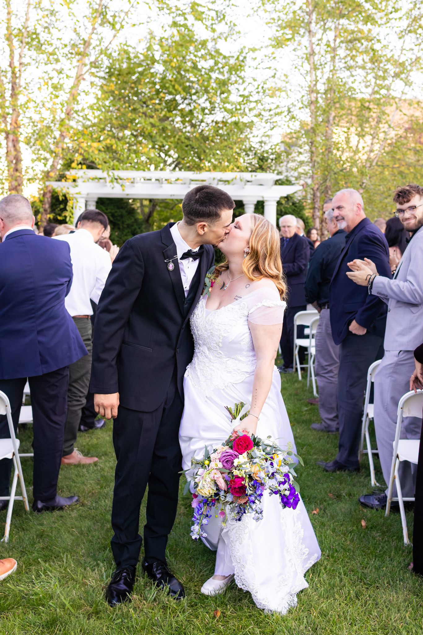Newlywed couple sharing a kiss at outdoor wedding ceremony on green lawn with white chairs and guests.