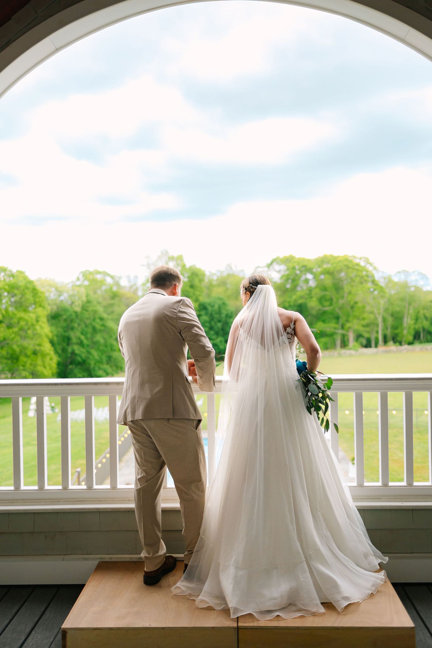 A wedding couple sits together on a white porch railing overlooking green trees on a sunny summer day.