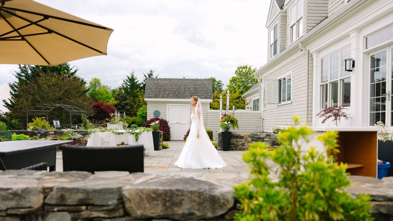 A wedding dress train trails across a stone patio with outdoor umbrellas and garden furniture in the background.