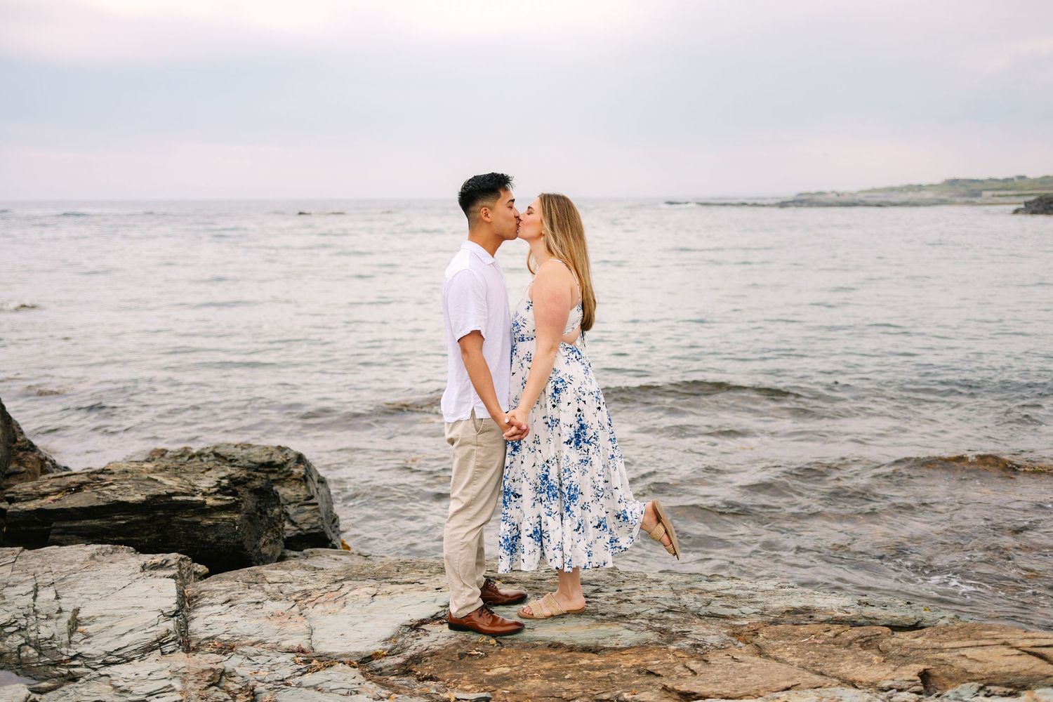 Newly engaged couple kissing on rocky shoreline in Newport Rhode Island, woman in floral dress, man in white shirt, ocean backdrop.