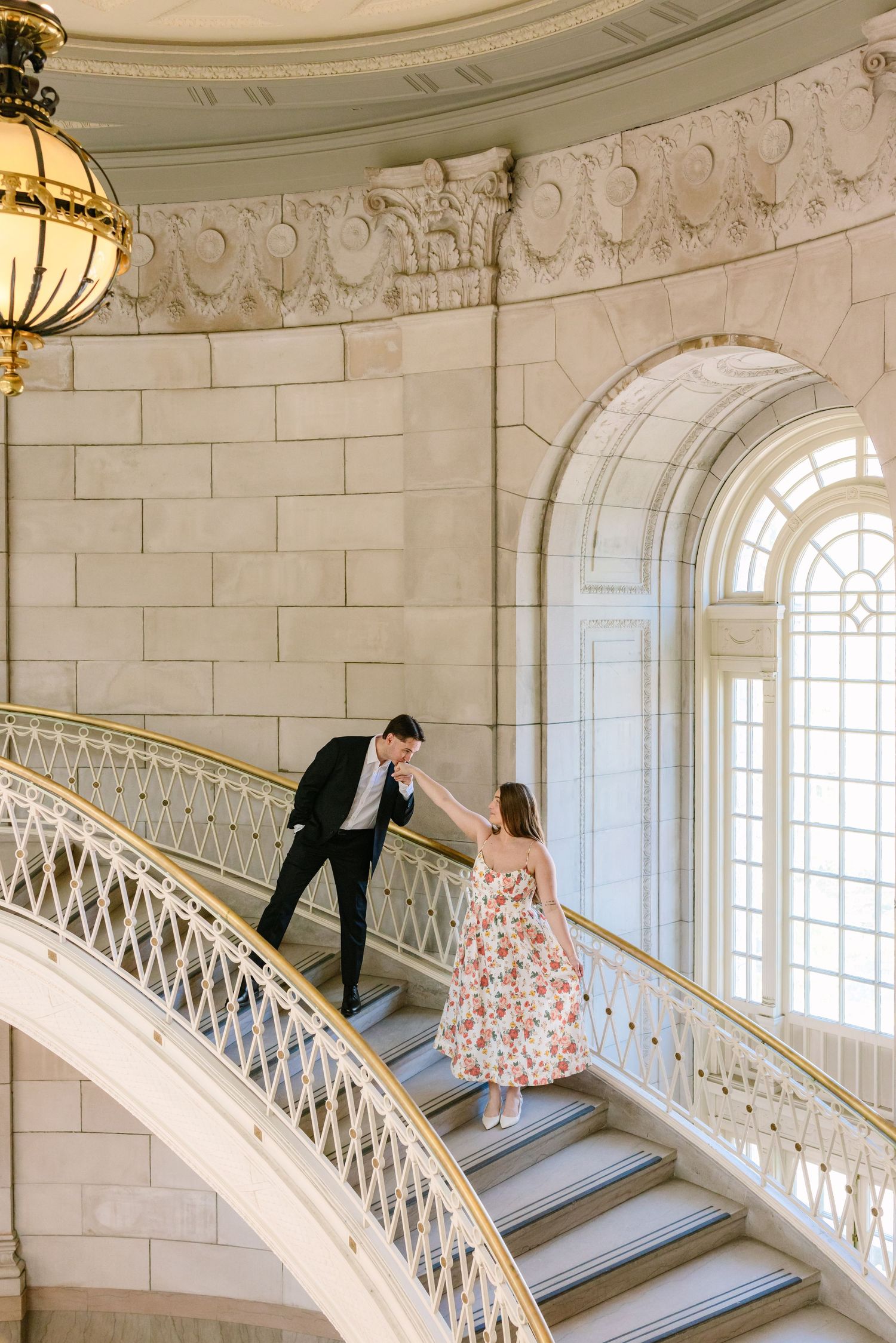 Couple holding hands on a sweeping curved staircase in a grand building.