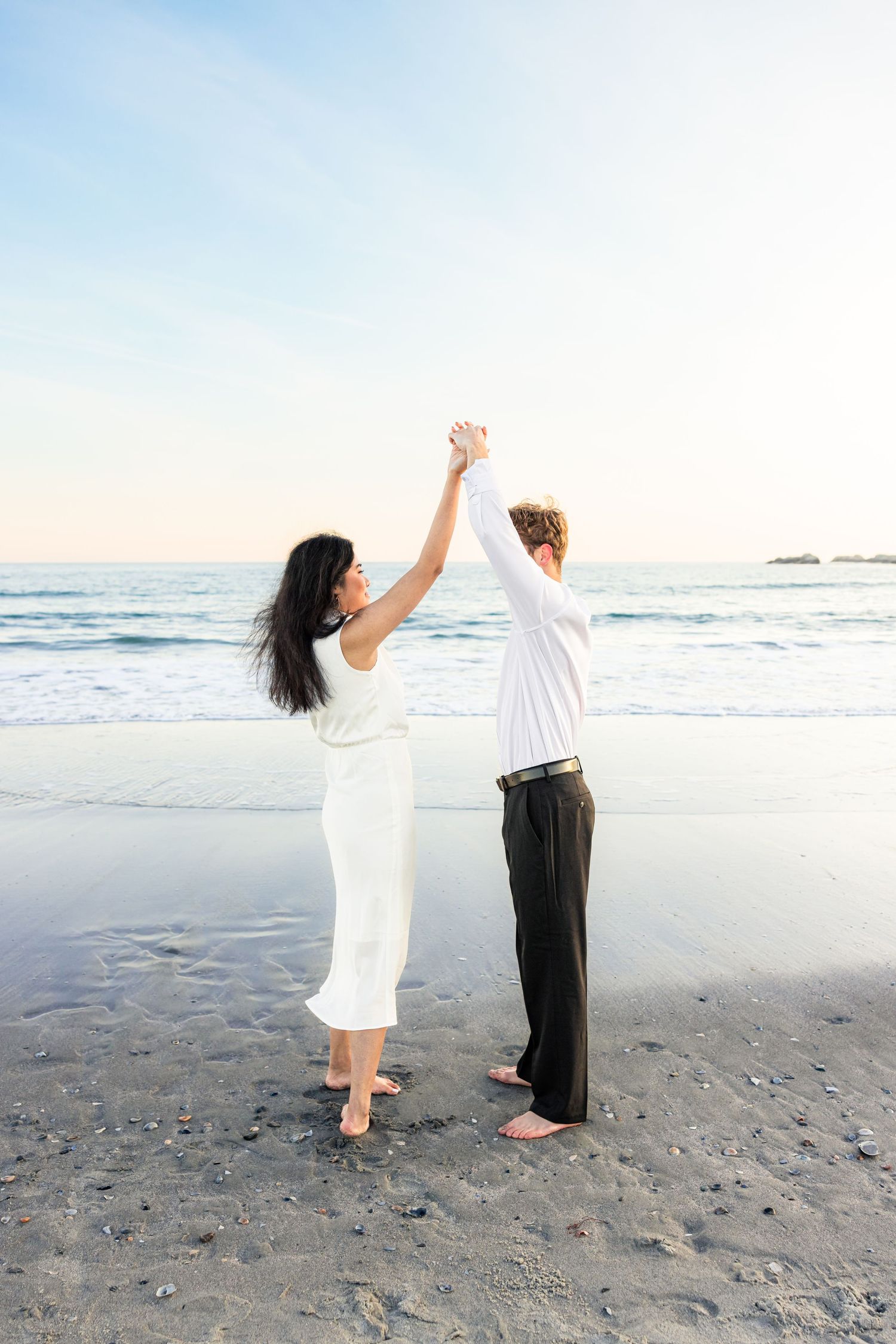 A couple dancing and holding hands on a serene beach at sunset with waves and blue sky in the background.