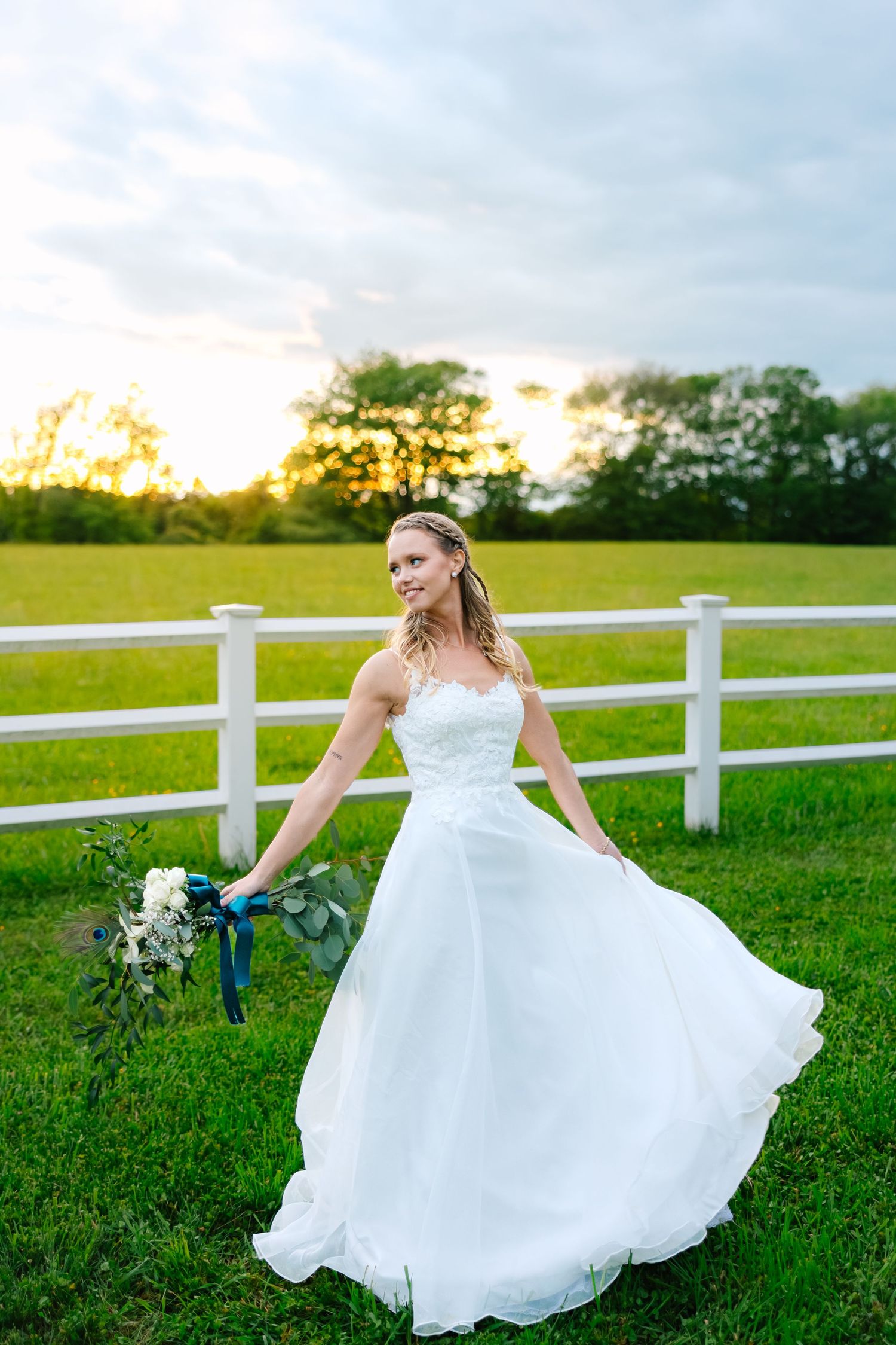 Elegant white wedding dress twirls on a grassy field at sunset with white fence and bouquet in countryside setting.