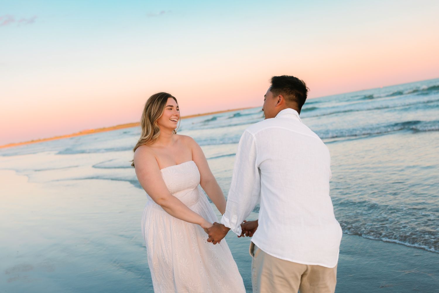 Couple holding hands on beach at sunset with pink and blue sky, woman in flowing white dress.