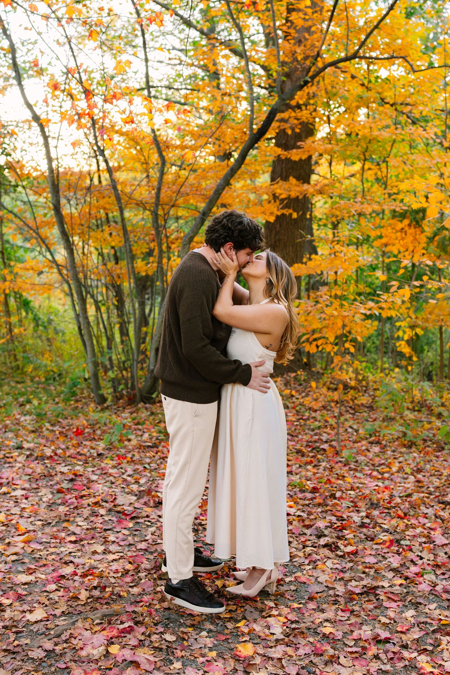A romantic autumn engagement photoshoot showing a couple embracing among vibrant fall foliage and scattered leaves.