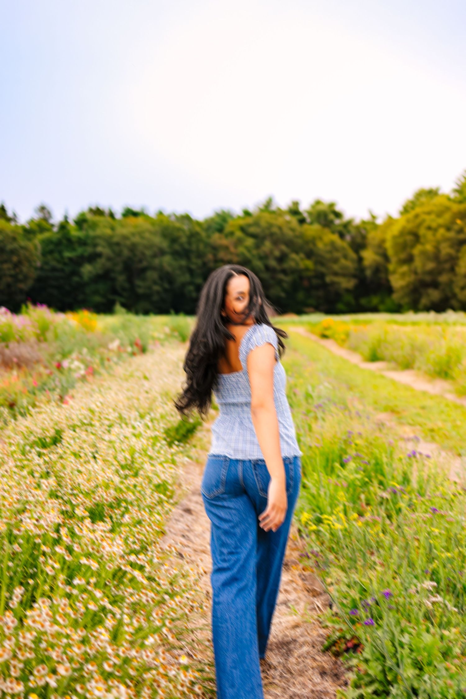 Young woman with long dark hair in blue top and jeans walking through rows of wildflower farm field.