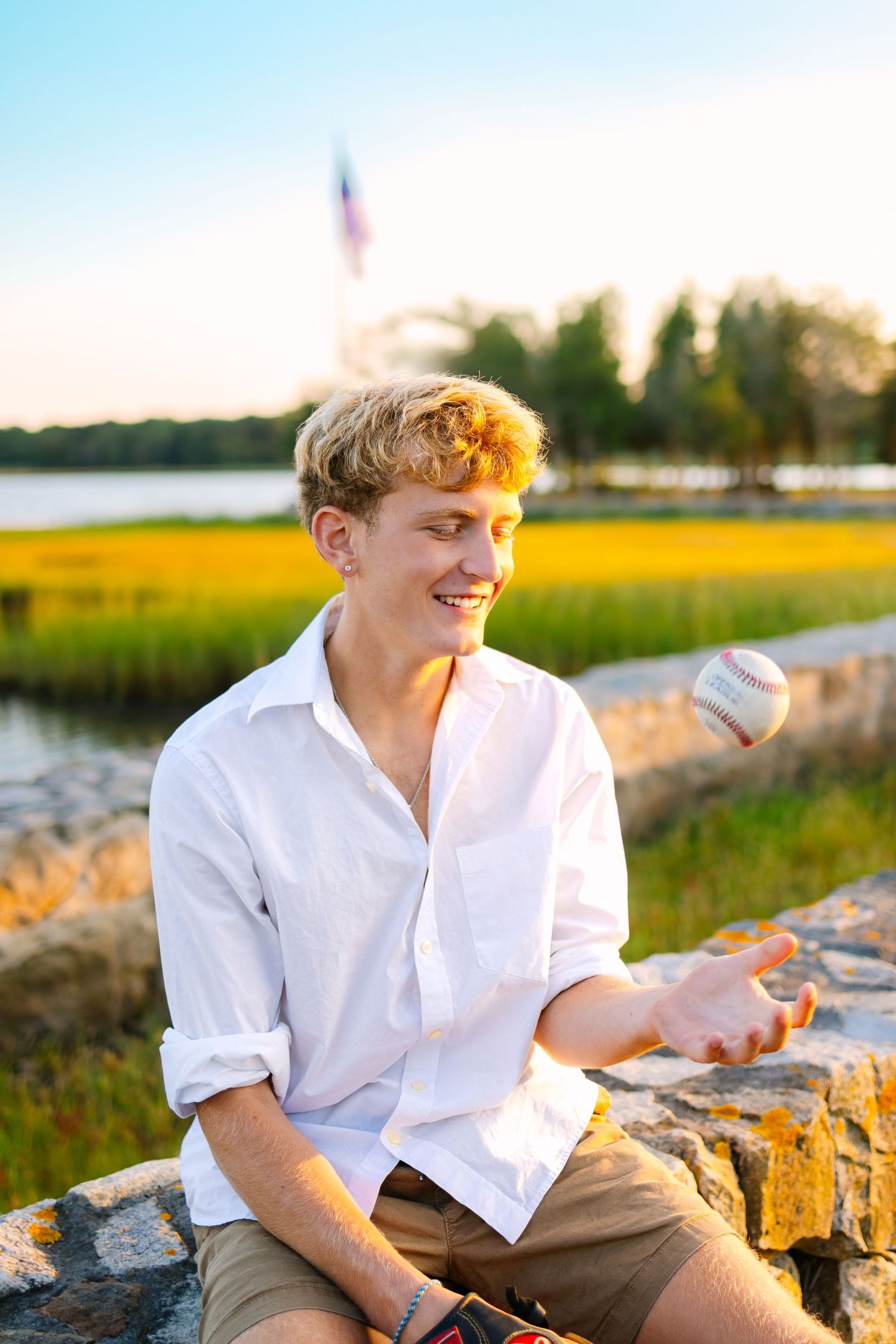 Happy teen boy in white shirt tosses baseball while sitting on stone wall with marsh and flag in background.