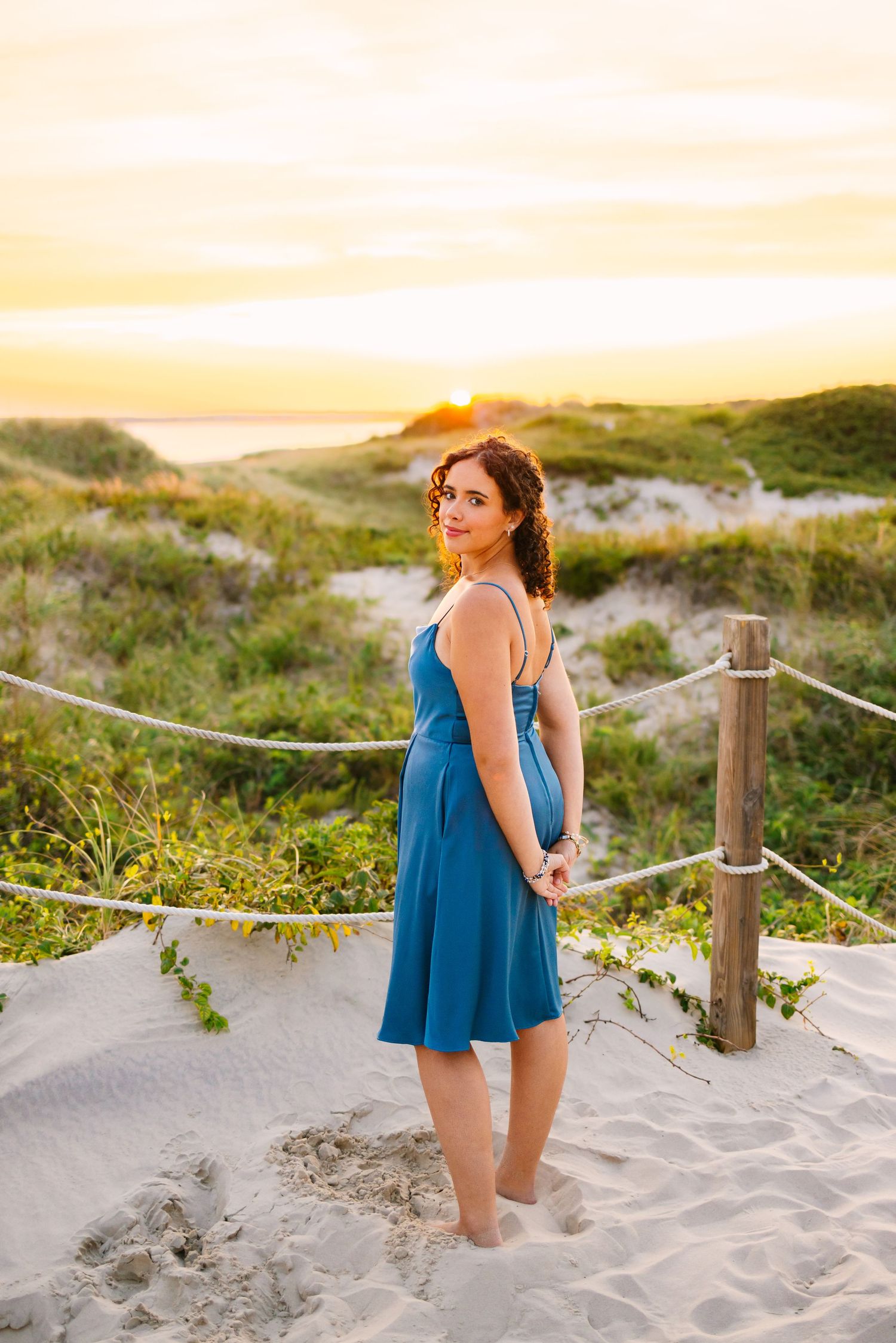 Woman in blue dress standing on sandy beach path near dunes at sunset.