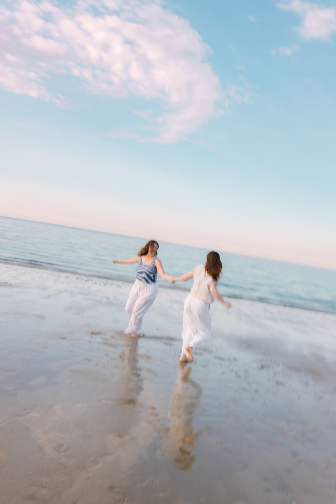 Two joyful women holding hands running toward ocean waves on a beach at sunset together.