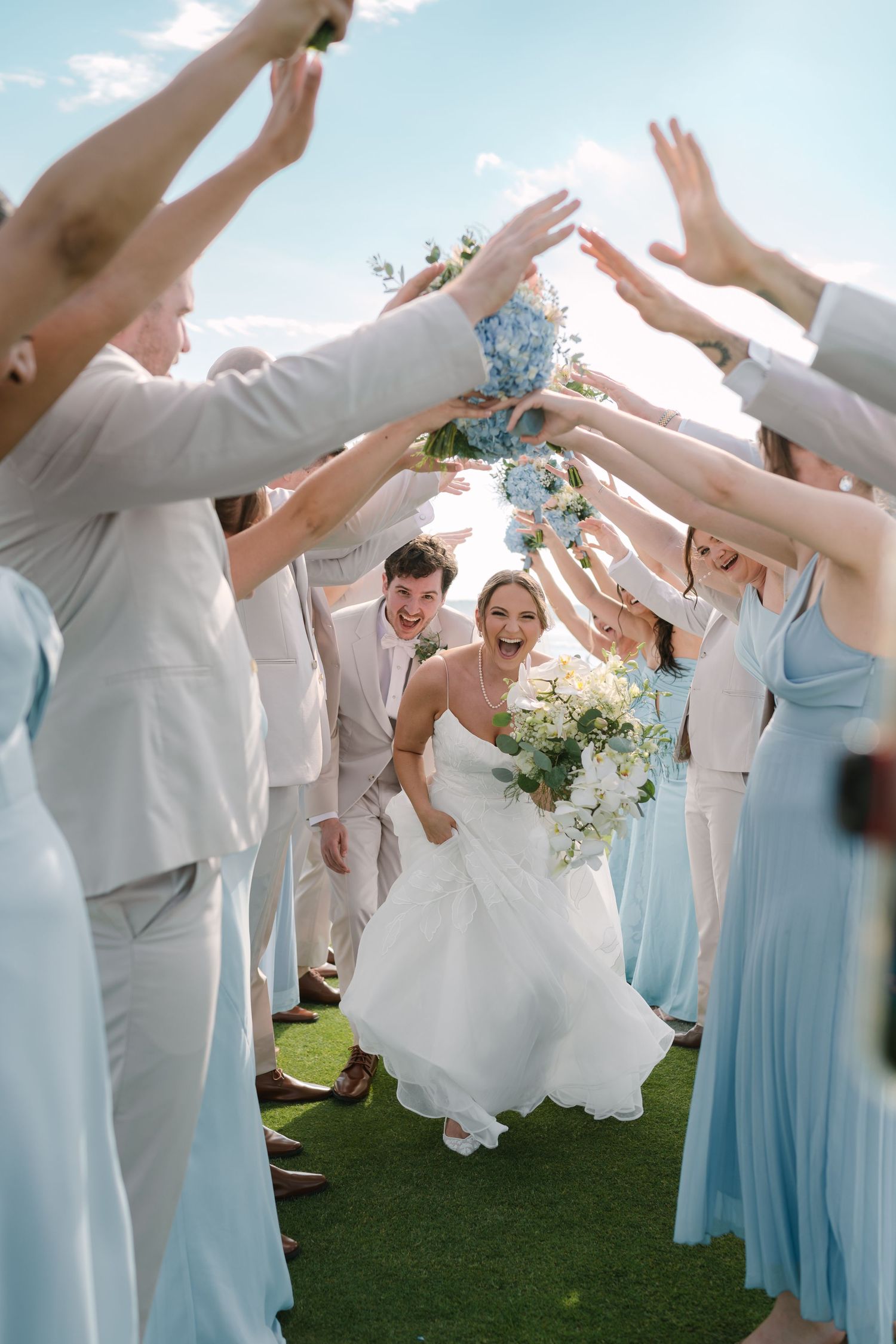 Bride and groom walking through an arch of raised arms formed by wedding party in blue and gray attire at Warwick Country Club in RI.