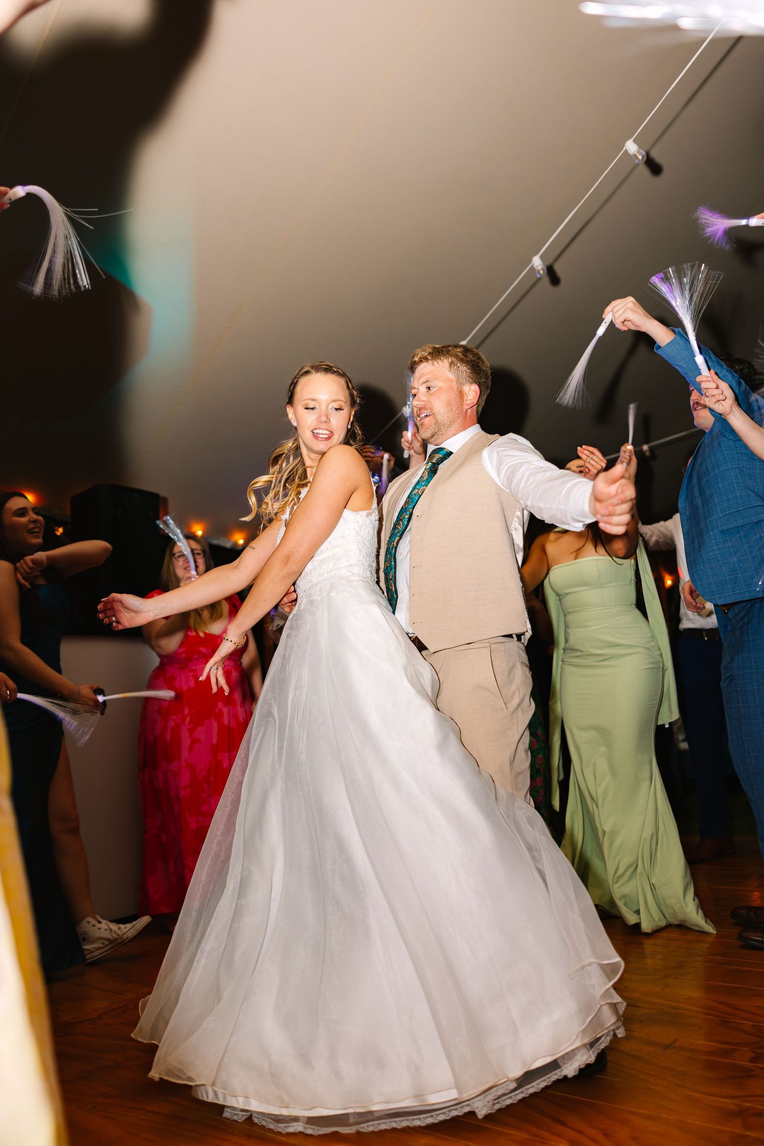 Bride in flowing white ballgown twirls on wooden dance floor during wedding reception with guests in green dresses.